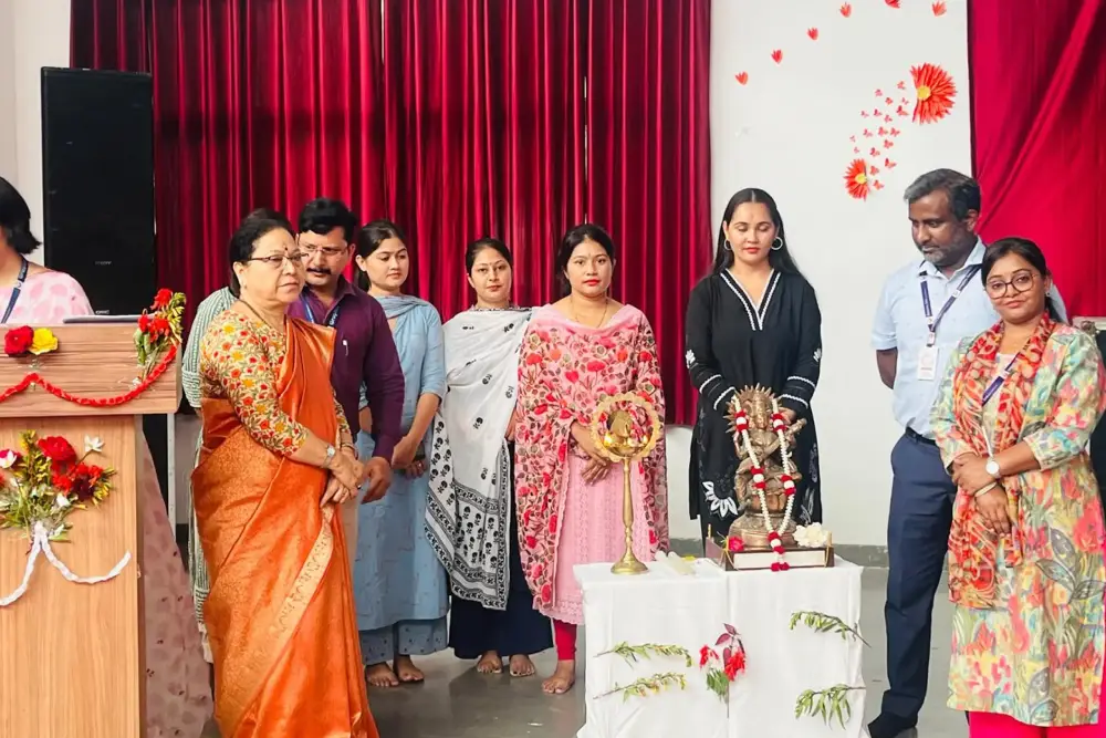 Group of people standing on a stage around a lit ceremonial lamp on a white-covered table during a cultural event, with red curtains and floral decorations in the background.