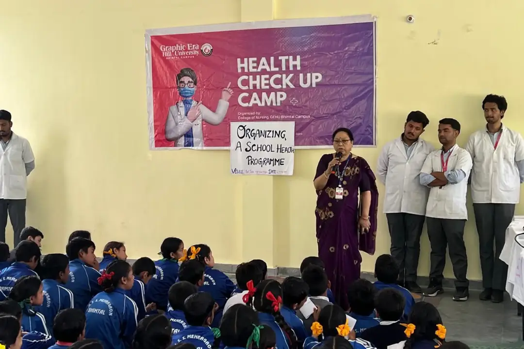 Speaker in purple sari addressing a large group of students during a health check-up camp, with banners behind her and medical staff nearby.
