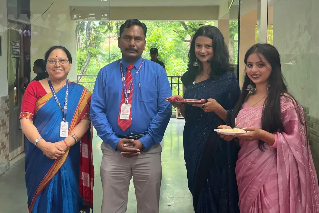 Four colleagues posing for a photo in a hallway; two women in sarees hold plates with snacks, a man in a blue shirt and red tie stands center, another woman in a dark blue saree on the right.