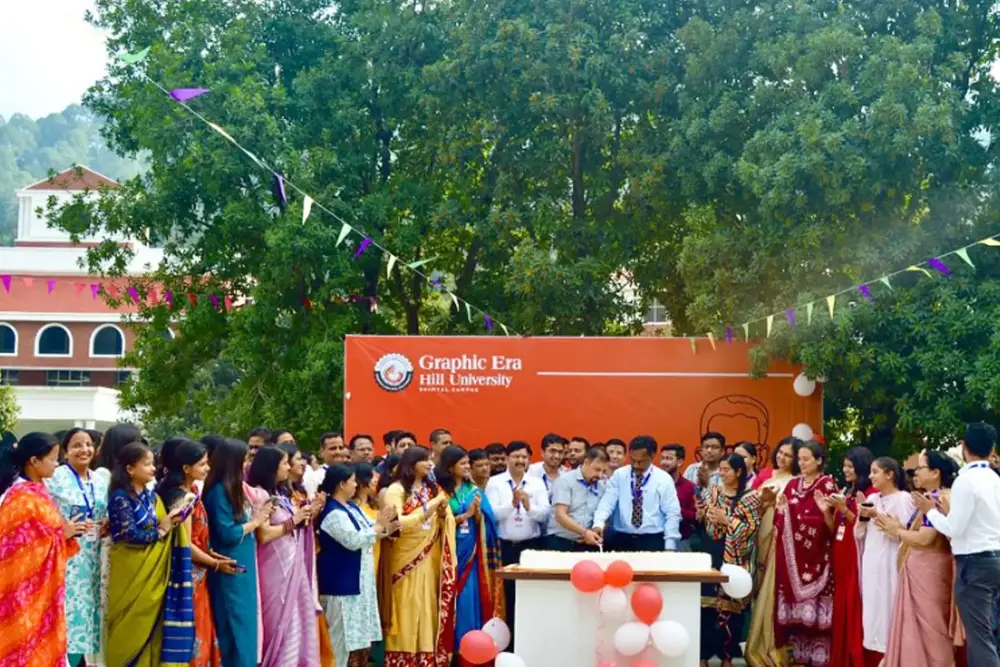 Group of students and staff in colorful attire at an outdoor university celebration, cutting a cake in front of an orange 'Graphic Era Hill University' banner.