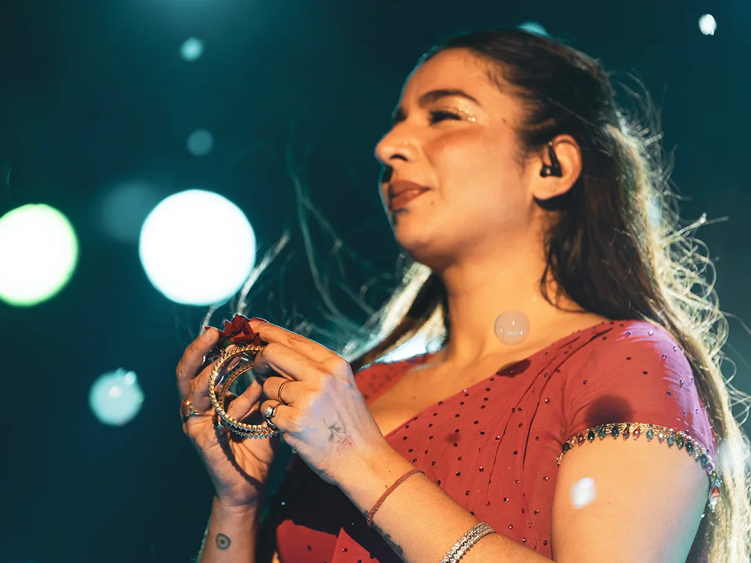 Singer on stage wearing a red dress with jewelry, holding bracelets near a mic, illuminated by stage lights.