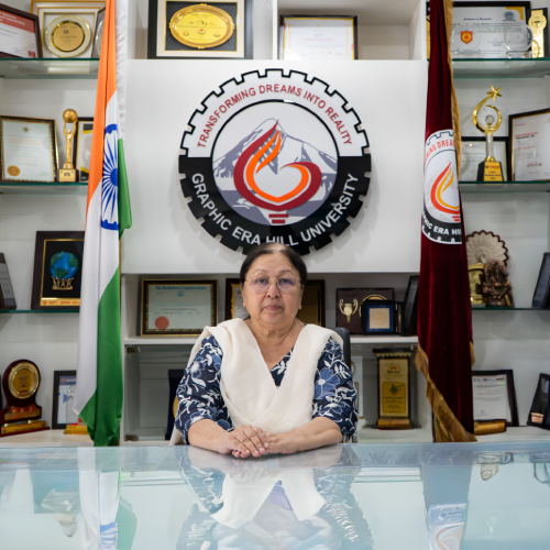 Woman seated at a glass desk in a university office, hands folded, with the Graphic Era Hill University emblem behind her and Indian flag to the left.